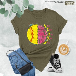 May include: Olive green t-shirt featuring a softball graphic with the text "ALL THE PRETTY GIRLS WALK LIKE THIS!". The shirt is paired with blue jeans, a black crossbody bag, and black high-top sneakers. A cup of coffee is also in the image.