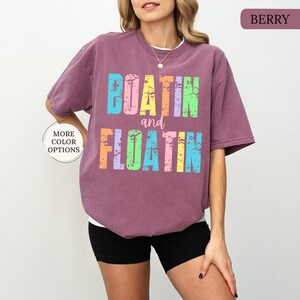 May include: Berry-coloured t-shirt with the words "BOATIN and FLOATIN" in colourful, distressed lettering. The shirt has a relaxed fit and short sleeves. A small button indicates more colour options.