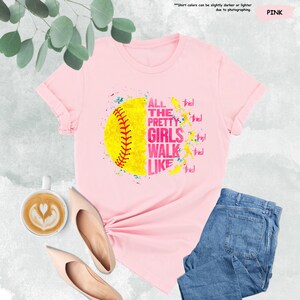 May include: Pink t-shirt featuring a yellow softball graphic with red stitching and the text "ALL THE PRETTY GIRLS WALK LIKE THIS!". The shirt is paired with blue jeans and nude-colored shoes. A cup of coffee with latte art is also visible.