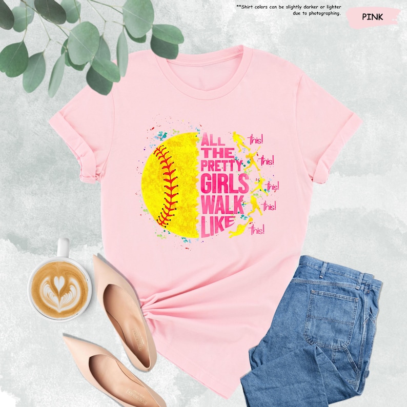 May include: Pink t-shirt featuring a yellow softball graphic with red stitching and the text "ALL THE PRETTY GIRLS WALK LIKE THIS!". The shirt is paired with blue jeans and nude-colored shoes. A cup of coffee with latte art is also visible.