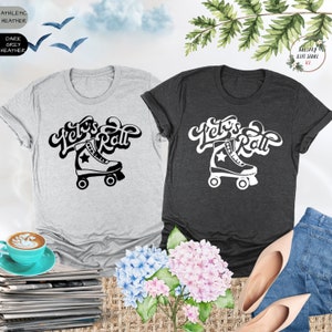 May include: Two t-shirts with a black and white graphic of a roller skate and the text "Let's Roll". The t-shirts are on a table with a cup of coffee, a stack of magazines, and flowers.