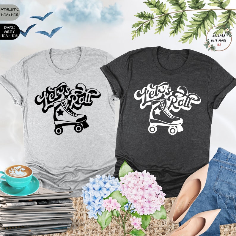 May include: Two t-shirts with a black and white graphic of a roller skate and the text "Let's Roll". The t-shirts are on a table with a cup of coffee, a stack of magazines, and flowers.
