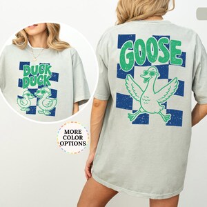 May include: A light gray short-sleeve t-shirt featuring a blue and green checkered pattern with a graphic of a goose wearing sunglasses and the text GOOSE on the back, and a smaller graphic of two ducks with the text DUCK DUCK on the front.