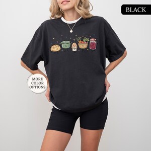 May include: A black crew neck t-shirt featuring a graphic print of a loaf of bread, a green pot, an egg, a basket of vegetables, and a jar of jam, all with smiling faces. The text "BLACK" appears in the top right corner and "MORE COLOR OPTIONS" in a circle.