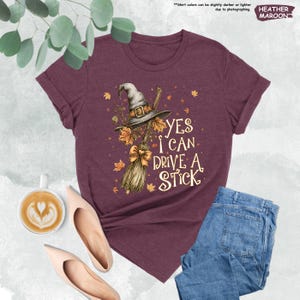 May include: A maroon short-sleeve t-shirt featuring a graphic of a witch hat and a broomstick with autumn leaves. The text reads, YES I CAN DRIVE A STICK. The design includes a small bow on the broom handle and scattered fall foliage accents.