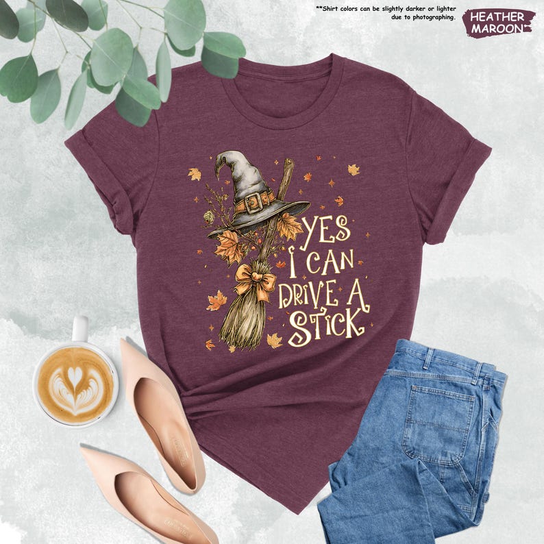 May include: A maroon short-sleeve t-shirt featuring a graphic of a witch hat and a broomstick with autumn leaves. The text reads, YES I CAN DRIVE A STICK. The design includes a small bow on the broom handle and scattered fall foliage accents.