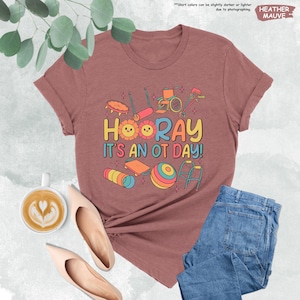 Peut inclure: T-shirt en mauve chin&eacute; avec un motif graphique d'outils d'ergoth&eacute;rapie et le texte "HOORAY IT'S AN OT DAY!"