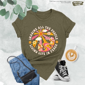 May include: A green t-shirt with a retro-style graphic of a sun, clouds, flowers, and mushrooms. The graphic is surrounded by the text "Imagine All The People Living Life In Peace".