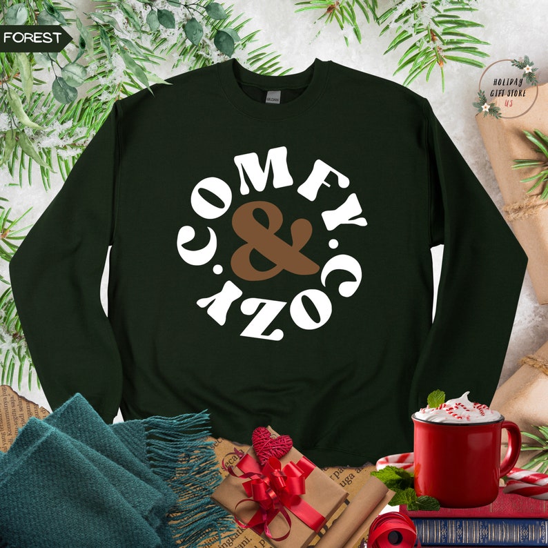 以下が含まれることがあります： レトロなフォントで「Comfy & Cozy」と書かれた白い円形のデザインが施された濃い緑色のスウェットシャツ。 「&」は茶色です。
