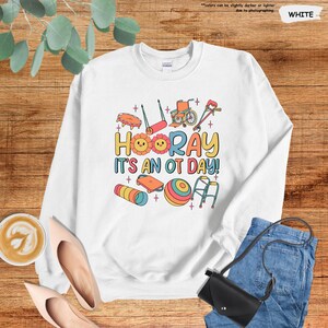 Peut inclure: Sweat-shirt blanc avec un motif graphique color&eacute; repr&eacute;sentant des outils d'ergoth&eacute;rapie et le texte "HOORAY IT'S AN OT DAY!"