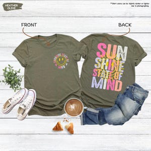 Pu&ograve; includere: T-shirt verde oliva melange con grafica "Sunshine State of Mind" sul davanti e sul retro. Il davanti presenta una faccina sorridente e testo, mentre il retro mostra le parole "Sun Shine State of Mind" a colori.