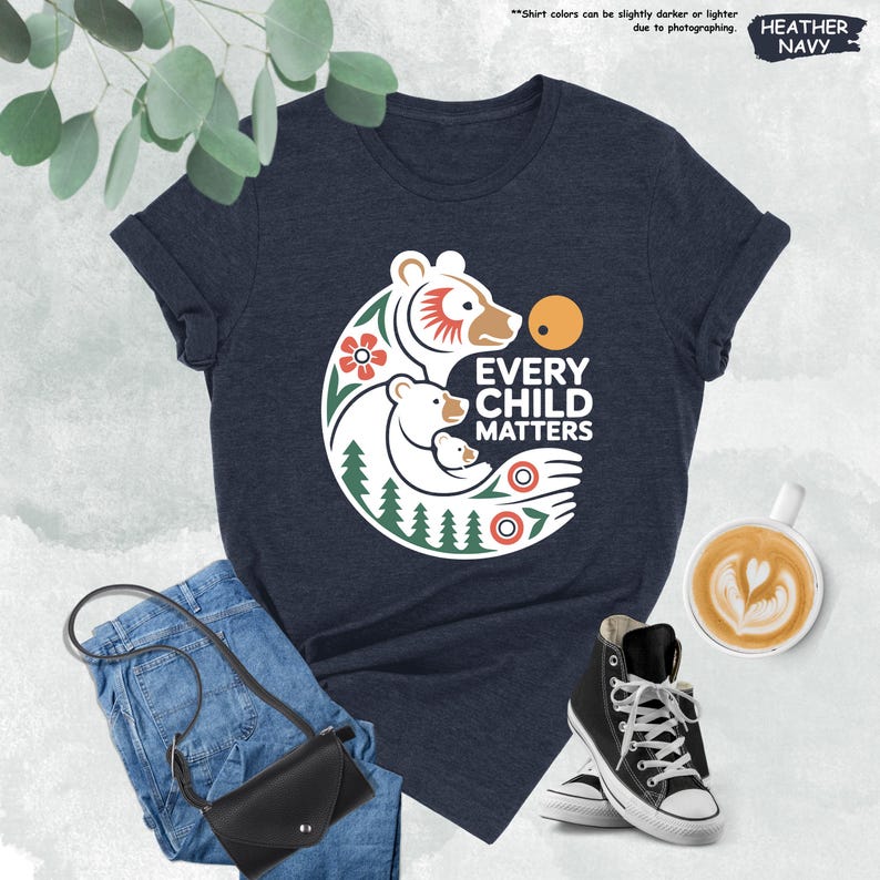May include: A navy blue t-shirt with the text "EVERY CHILD MATTERS" and a graphic of a bear family. The design includes a bear, a cub, flowers, and trees. The shirt is paired with blue jeans, a black purse, and black high-top sneakers.