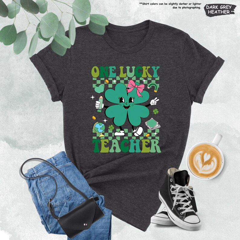 May include: Dark heather grey t-shirt with a green four-leaf clover graphic. The clover has a smiling face, a pink bow, and is holding a pencil. The text "One Lucky Teacher" is printed above the clover.