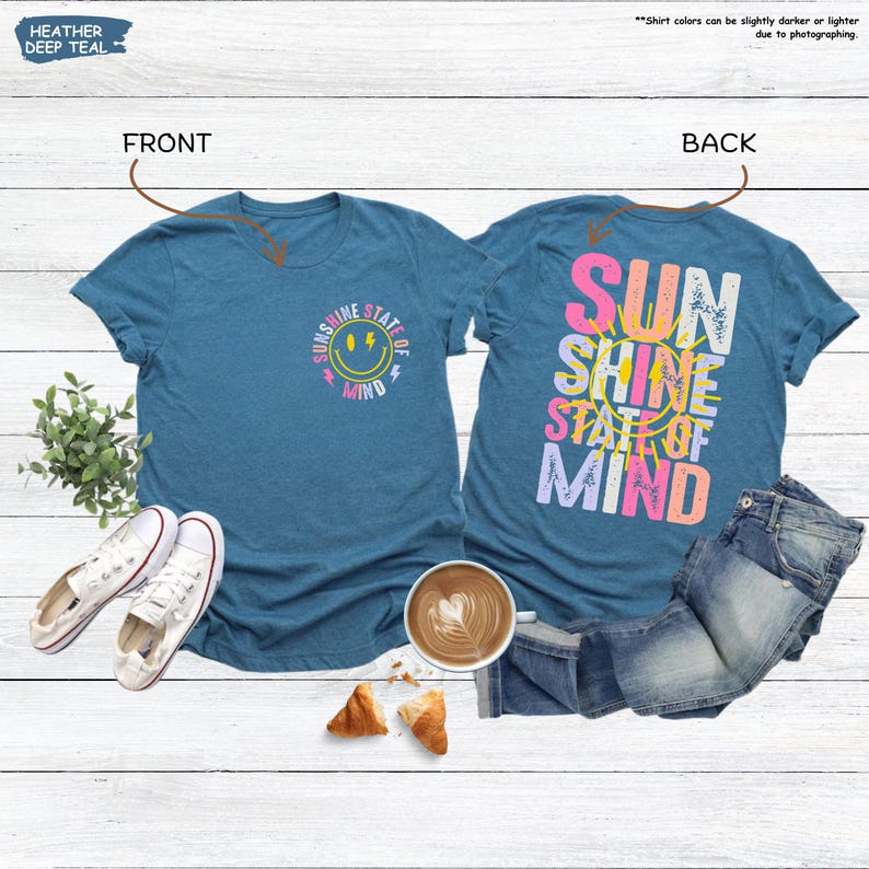 Pu&ograve; includere: T-shirt color verde acqua con la scritta "Sunshine State of Mind" sul davanti e sul retro. Il davanti presenta una faccina sorridente e il retro le parole in un design colorato. Scarpe da ginnastica bianche e jeans accanto alle magliette.