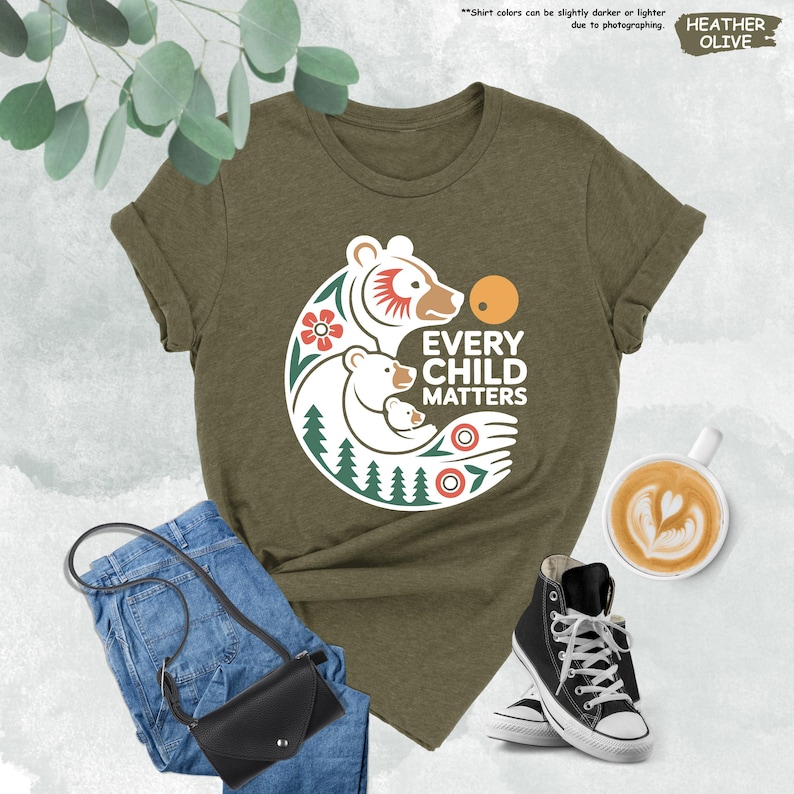 May include: Heather olive green t-shirt with a graphic of three bears, a flower, and the text "EVERY CHILD MATTERS". The shirt is styled with blue jeans, a black purse, and black high-top sneakers.