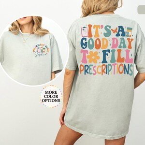 May include: A light green t-shirt with the text "IT'S A GOOD DAY TO FILL PRESCRIPTIONS" in colourful, retro-style lettering on the back. The front features a pharmacy tech design with the name "Sophia".