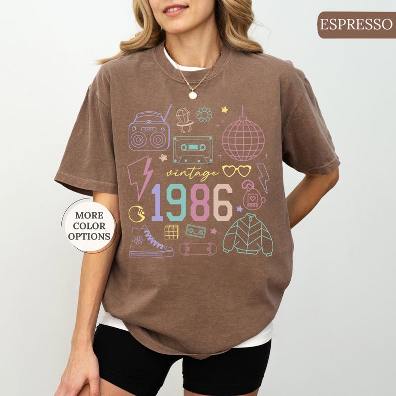 May include: Espresso-colored t-shirt with a vintage 1986 graphic. The design includes a boombox, cassette tape, disco ball, and other retro icons in pastel colors. The shirt has short sleeves and a crew neck.