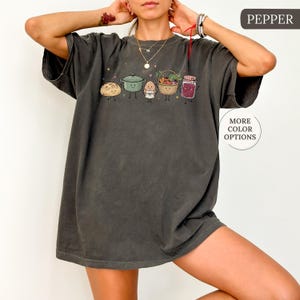 May include: A charcoal gray short-sleeve t-shirt featuring a graphic print of five anthropomorphic food items including a loaf of bread, a green pot, an egg, a basket of vegetables, and a jar of jam. The text PEPPER appears in the top right corner.