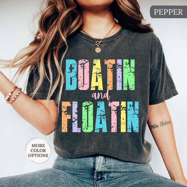 May include: Dark grey t-shirt featuring the text "BOATIN and FLOATIN" in distressed, multi-coloured lettering. The shirt has a vintage appearance and is a casual style.