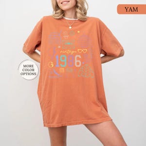 May include: A yam-colored t-shirt with a retro design. The shirt features the text "vintage 1986" with colorful graphics of a cassette tape, disco ball, and other 80s-themed icons. The shirt has short sleeves and a relaxed fit.