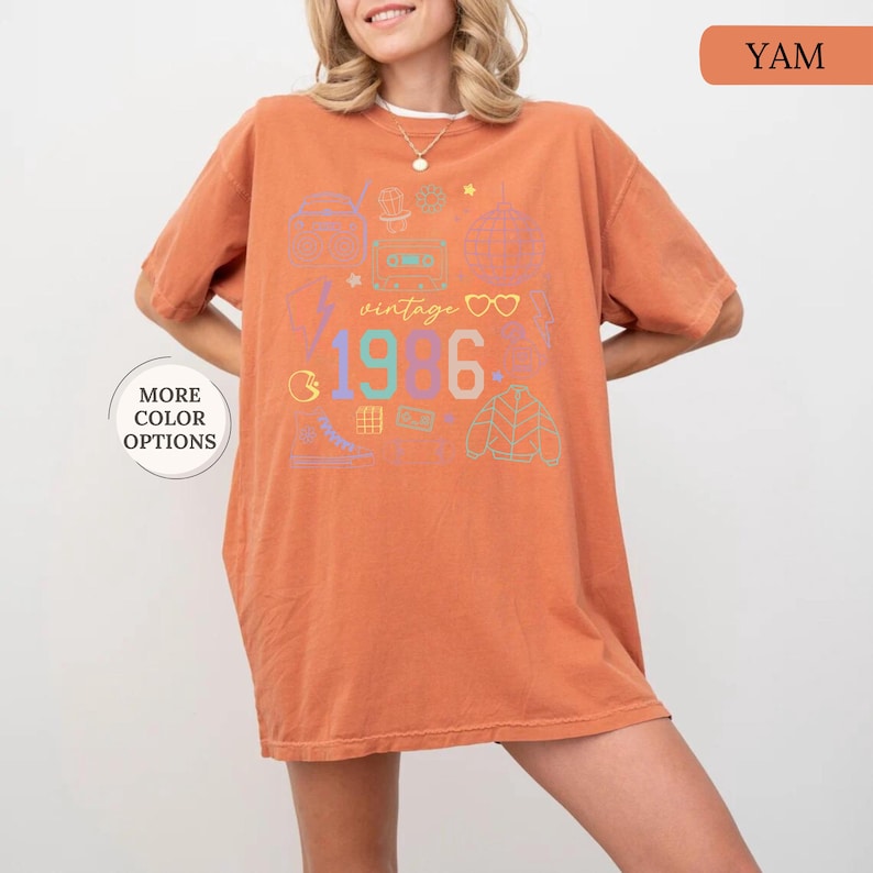 May include: A yam-colored t-shirt with a retro design. The shirt features the text "vintage 1986" with colorful graphics of a cassette tape, disco ball, and other 80s-themed icons. The shirt has short sleeves and a relaxed fit.