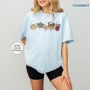 May include: A light blue t-shirt featuring a graphic print of a loaf of bread, a green pot, an egg, a basket of vegetables, and a jar of jam, all with smiling faces. The text CHAMBRAY appears in the top right corner and MORE COLOR OPTIONS in a circle.
