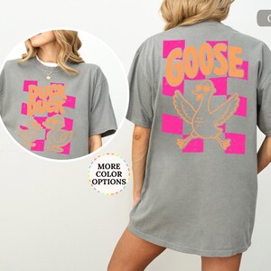 May include: A gray t-shirt featuring a back graphic of a goose wearing sunglasses over a pink and orange checkered pattern with the text GOOSE. The front chest area displays a smaller graphic of two ducks with the text DUCK DUCK.