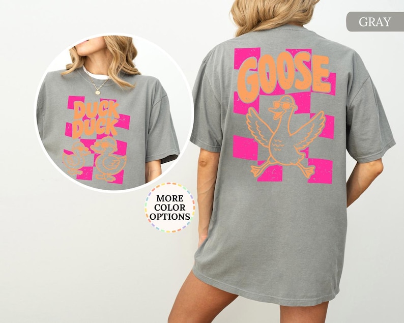 May include: A gray t-shirt featuring a back graphic of a goose wearing sunglasses over a pink and orange checkered pattern with the text GOOSE. The front chest area displays a smaller graphic of two ducks with the text DUCK DUCK.
