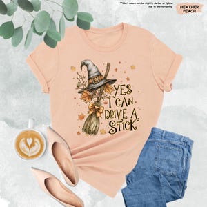 May include: A heather peach t-shirt featuring a graphic of a witch hat and a broomstick with autumn leaves. The text reads, Yes I Can Drive A Stick. The design includes a small bow on the broom handle and scattered fall foliage elements.