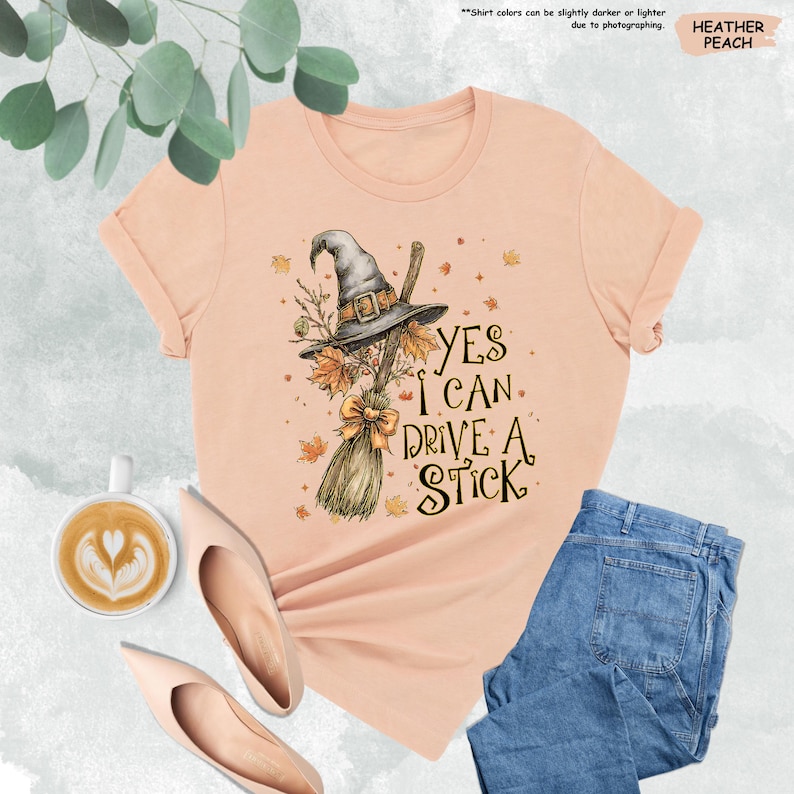 May include: A heather peach t-shirt featuring a graphic of a witch hat and a broomstick with autumn leaves. The text reads, Yes I Can Drive A Stick. The design includes a small bow on the broom handle and scattered fall foliage elements.