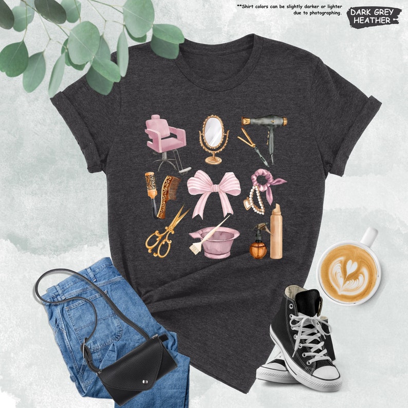 Op de afbeelding: Donkergrijs t-shirt met een grafisch ontwerp van verschillende kapsalon gereedschappen en accessoires, waaronder een roze kapsalonstoel, een f&ouml;hn, een schaar, een haarborstel, een haarlakfles en een roze strik. De tekst "Shirtkleuren kunnen door de fotografie iets donkerder of lichter zijn. DONKERGRIJ MELANGE" staat bovenaan de afbeelding.
