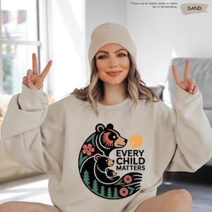 May include: Sand-colored sweatshirt with a graphic of a bear family, flowers, and the text "EVERY CHILD MATTERS." The sweatshirt is paired with a matching beanie. The image also includes the text "colors can be slightly darker or lighter due to photographing."