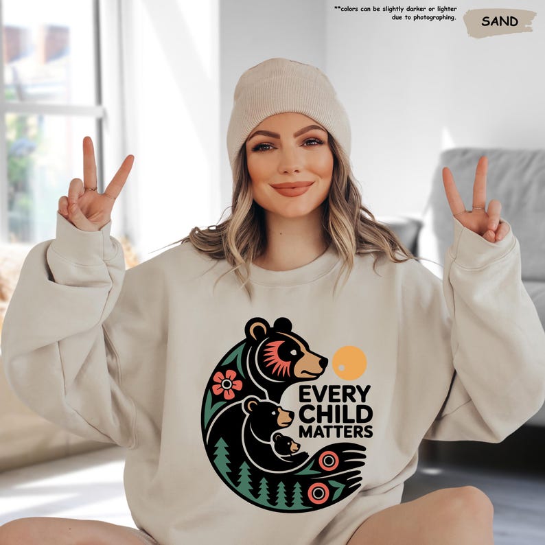 May include: Sand-colored sweatshirt with a graphic of a bear family, flowers, and the text "EVERY CHILD MATTERS." The sweatshirt is paired with a matching beanie. The image also includes the text "colors can be slightly darker or lighter due to photographing."