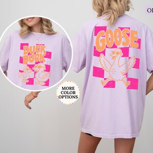 May include: An orchid-colored t-shirt featuring a graphic on the back of a goose wearing sunglasses above a pink checkered pattern with the text GOOSE. The front shows two ducks wearing sunglasses with the text DUCK DUCK over a pink checkered pattern.