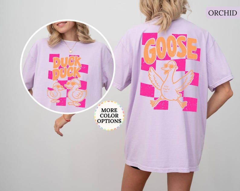 May include: An orchid-colored t-shirt featuring a graphic on the back of a goose wearing sunglasses above a pink checkered pattern with the text GOOSE. The front shows two ducks wearing sunglasses with the text DUCK DUCK over a pink checkered pattern.