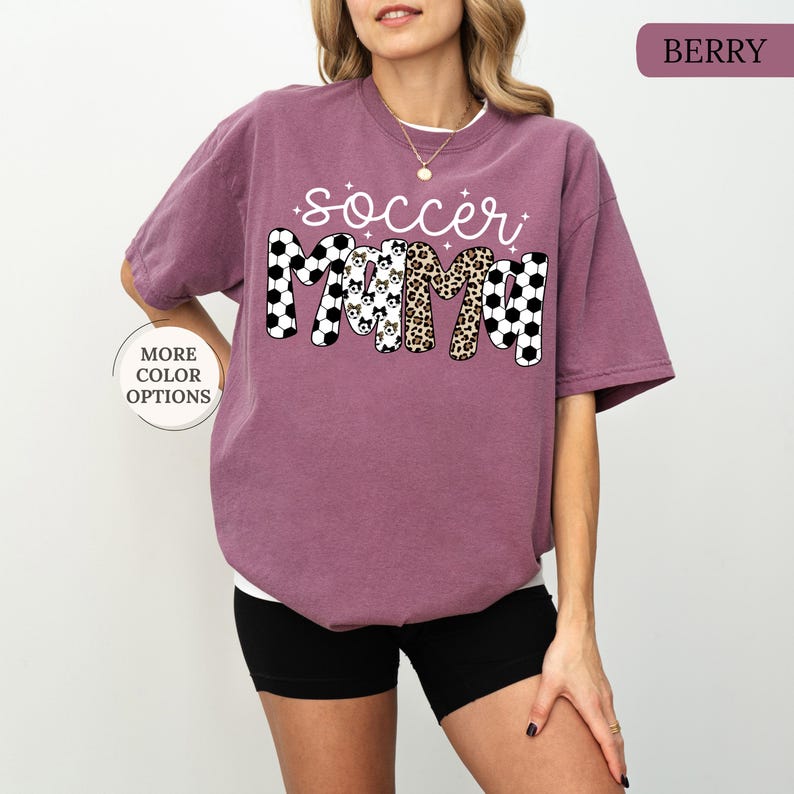 May include: Berry-coloured t-shirt featuring the text "soccer mama". The letters are decorated with soccer ball patterns, animal prints, and dog faces. The shirt has short sleeves and a relaxed fit. The colour is described as "Berry".