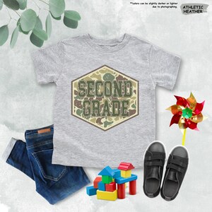 May include: A grey t-shirt with the words "SECOND GRADE" in a camouflage design. The image also shows a pair of dark blue jeans, black trainers, a colourful pinwheel, and wooden building blocks.