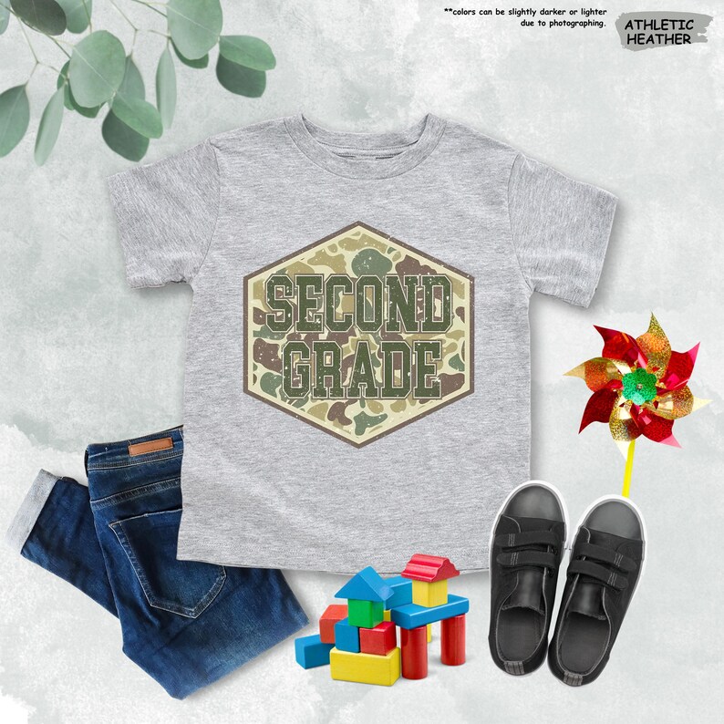 May include: A grey t-shirt with the words "SECOND GRADE" in a camouflage design. The image also shows a pair of dark blue jeans, black trainers, a colourful pinwheel, and wooden building blocks.