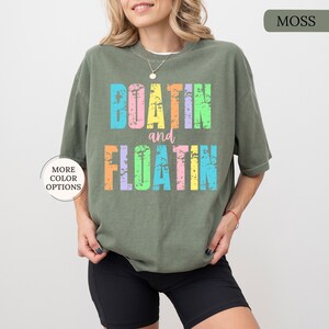 May include: A moss green t-shirt with the words "BOATIN and FLOATIN" in colourful, distressed lettering. The shirt has a relaxed fit and short sleeves. The words are in pastel colours, including blue, pink, yellow, and orange.