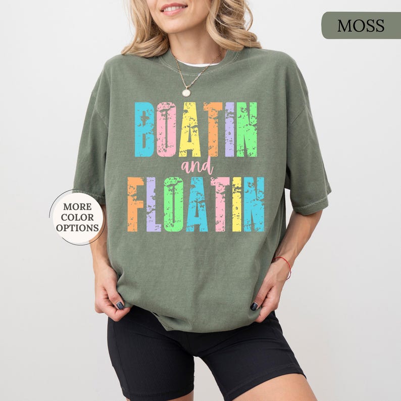 May include: A moss green t-shirt with the words "BOATIN and FLOATIN" in colourful, distressed lettering. The shirt has a relaxed fit and short sleeves. The words are in pastel colours, including blue, pink, yellow, and orange.