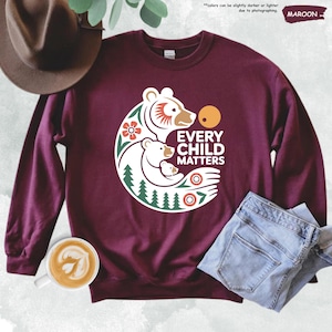 May include: Maroon sweatshirt with a white graphic of three bears, a flower, and the text "EVERY CHILD MATTERS." The design includes a sun, trees, and other decorative elements. The sweatshirt is paired with blue jeans and a brown hat.