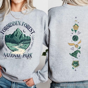May include: Gray sweatshirt with a green and white graphic of a forest scene with the text "Forbidden Forest National Park Est. 500TLAND 880 AD". The back of the sweatshirt has a vertical graphic of a potion bottle, a golden snitch, a wizard's hat, and a crest.
