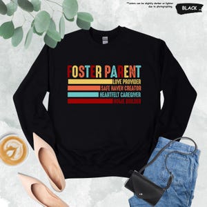 May include: Black sweatshirt with the text "FOSTER PARENT" in a retro style font. The text is layered over colorful stripes. The text below the title reads "LOVE PROVIDER", "SAFE HAVEN CREATOR", "HEARTFELT CAREGIVER", and "HOME BUILDER".