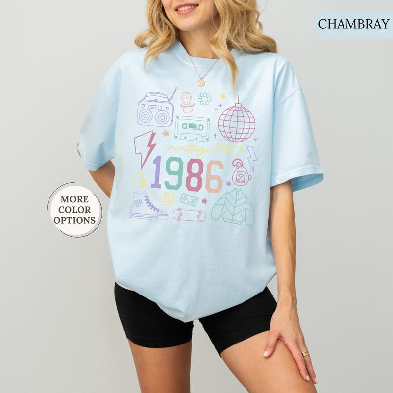 May include: Light blue t-shirt with a vintage 1986 graphic. The design includes a boombox, cassette tape, disco ball, and other retro icons in pastel colors. The shirt has short sleeves and a crew neck.