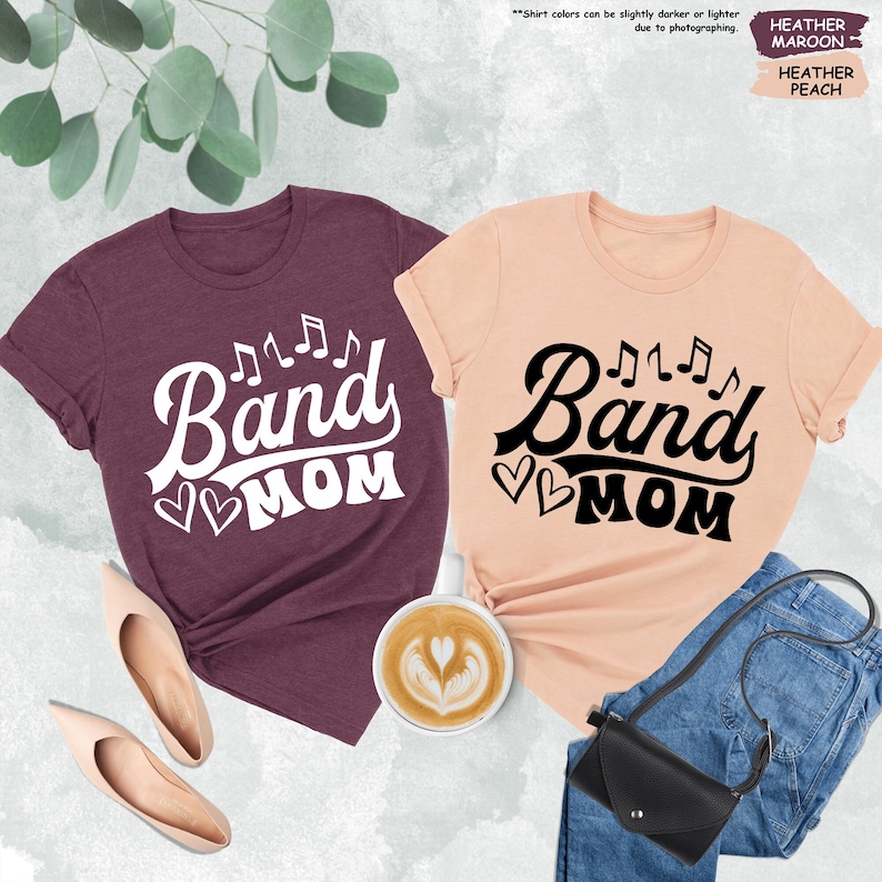 Może przedstawiać: Dwie koszulki z napisem "Band Mom" i nutami. Jedna koszulka jest w kolorze brązowym melanż, a druga w kolorze brzoskwiniowym melanż.
