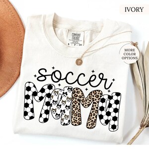May include: An ivory-coloured t-shirt with the words "soccer mama" in black script and block letters. The word "mama" features soccer ball patterns, leopard print, and soccer ball patterns with bows. A gold necklace is draped on the shirt.