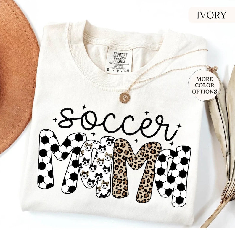 May include: An ivory-coloured t-shirt with the words "soccer mama" in black script and block letters. The word "mama" features soccer ball patterns, leopard print, and soccer ball patterns with bows. A gold necklace is draped on the shirt.