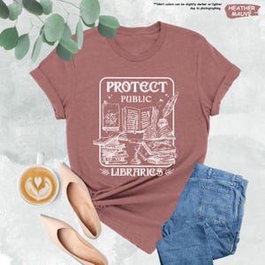 May include: A heather mauve t-shirt with a white graphic design that says "Protect Public Libraries" with an illustration of books, a quill pen, and a bottle of ink.