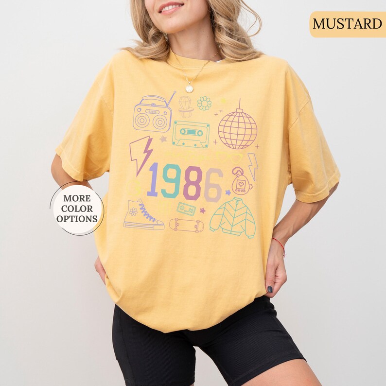 May include: Mustard-colored t-shirt with a retro 1986 graphic. The design includes a boombox, cassette tape, disco ball, and other 80s-themed illustrations in pastel colors. The shirt is a relaxed fit.