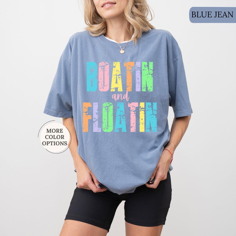 May include: A blue jean coloured t-shirt with the words "BOATIN and FLOATIN" in colourful, distressed lettering. The shirt has a relaxed fit and is worn with black shorts. The words "MORE COLOR OPTIONS" are also visible.